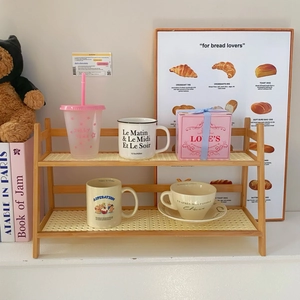 Detail of a 2-tier cream rattan desktop organizer shelf holding mugs and gift boxes
