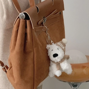 Close-up of white plush dog charm with brown knit hat and scarf on a suede bag