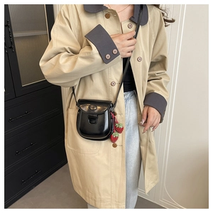 Détail d'un sac besace noir brillant avec breloques fraises rouges sur un trench beige
