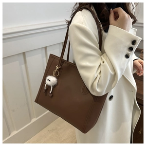 Brown leather tote bag with a plush white doll pendant shown from a side profile