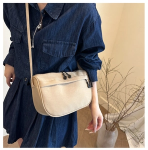 Close-up of beige canvas crossbody bag with dual zippers and logo detail on blue dress