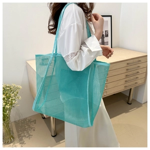 Detail of a large teal mesh tote bag carried by a model in a minimalist indoor setting