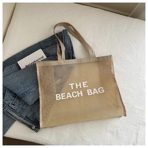 Borsa da spiaggia in rete beige distesa su sfondo in tessuto bianco con scritte grandi