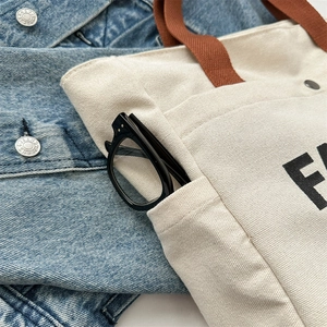 Cream canvas tote bag with brown handles, large printed letters, and side storage pocket