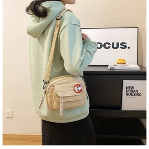 Detail view of beige canvas crossbody bag featuring a cartoon badge and woven rope zipper pull
