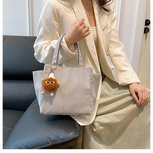Gray corduroy tote bag with side pockets and a cute plush charm on a black leather sofa