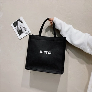 Detail of a black canvas tote bag with white merci text held against a beige wall