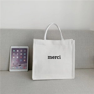 Detail showing the white canvas tote bag with 'merci' print and iPad for scale