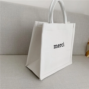 Minimalist white canvas tote bag with black merci print on a soft grey sofa texture