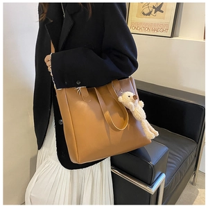 Brown leather tote bag with plush bear charm worn over shoulder in elegant city style