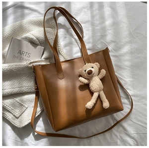 Brown faux leather tote bag with plush bear charm on white fabric background