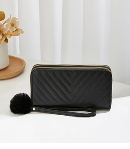 Close-up of black quilted long purse with dual gold zippers and fluffy pompom charm