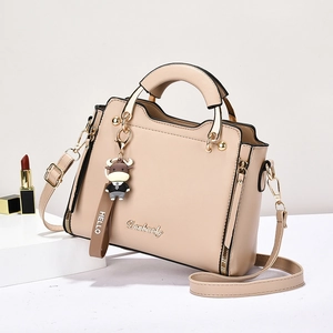 Minimalist beige crossbody handbag with top handle and cartoon pendant on white stand