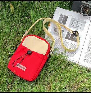 Close-up of red and beige crossbody bag with drawstring pocket and GUANG-YU logo on grass