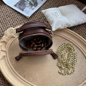 Mini round brown crossbody bag with gold chain strap and zipper detail shown on a tray