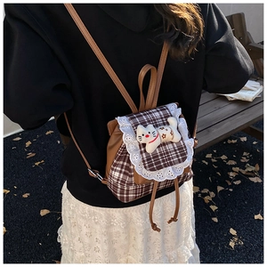 Lace-trimmed plaid backpack with white kitten plush toy on an outdoor wooden bench