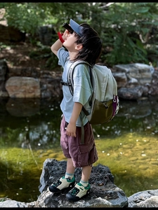 Kid using binoculars wearing blue t-shirt and hiking backpack by a mountain stream