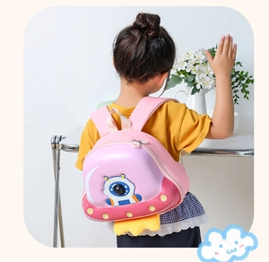 Pink cartoon UFO astronaut shell backpack for kids shown on a model in a home setting