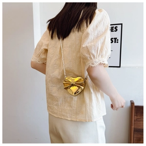 Close-up of golden heart-shaped crossbody bag with bow detail and pearl chain on a model