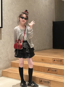 Fillette en gilet gris et jupe noire à volants avec un mini sac à bandoulière rouge matelassé