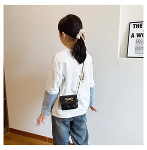 Detail of a black quilted crossbody bag with a gold bow and leather chain strap on a child