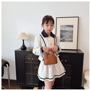 Girl in white puff-sleeve dress carrying a brown faux leather shell bag in a home setting
