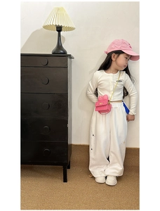 Girl in white cardigan and pink bow crossbody bag standing by a cabinet in home setting
