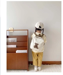 Toddler wearing a cream canvas bear backpack with adjustable straps in a warm nursery setting