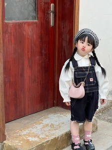 Kid wearing white blouse with black denim overalls and a pink chain saddle bag outdoors