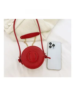 Mini round red PU crossbody bag with golden chain shown next to smartphone for size scale