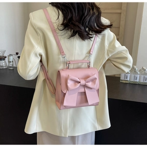 Pink matte leather backpack with large bow detail shown on model in a cozy cafe setting