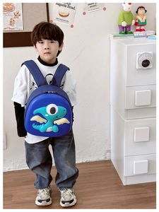 Blue dinosaur hardshell toddler backpack shown on a boy in a minimalist indoor setting