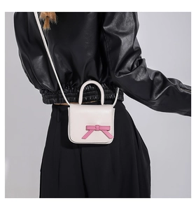 Detailed view of white mini crossbody bag with pink bow accent worn with black jacket