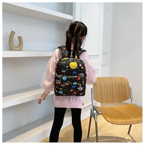 Girl wearing black cartoon space backpack with planets and rockets in a bright room