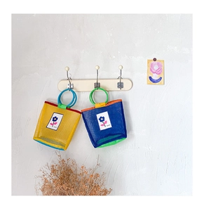 Détail de sacs en maille colorés avec anses rondes et logo floral sur un mur texturé