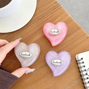 Glittery heart phone grip stands in pink, purple, and pearl white on a wooden desk