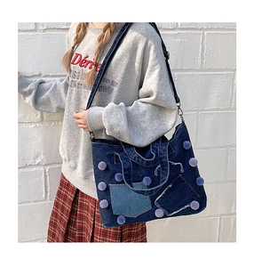 Dettaglio borsa tote in denim blu con pompon viola e tasche patchwork su muro di mattoni