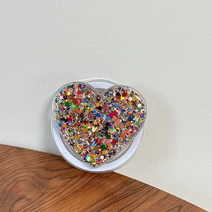 Close-up of heart-shaped phone grip filled with colorful sequins on white base