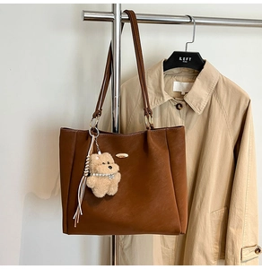 Detail of a brown leather tote bag with a golden metal logo and a plush bear pendant