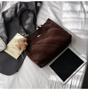 Brown leather tote bag featuring a gold oval plaque and a plush bear pendant with pearl chain