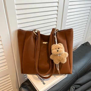 Brown matte leather tote bag with teddy bear charm on a book against window blinds