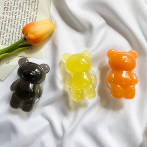Three 3D resin bear phone grips in translucent grey, yellow, and orange on white fabric