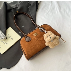 Detail der braunen Wildleder-Schultertasche mit Goldlogo und Plüschbär auf weißem Stoff