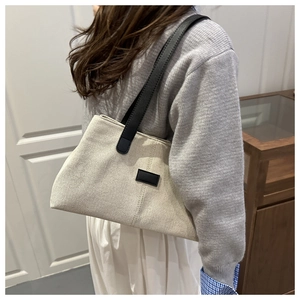Detail of beige tote bag with black straps and side patch shown on shoulder in interior setting