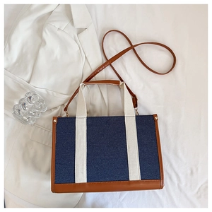 Flat lay of blue denim tote bag with white handles and brown leather trim on white fabric