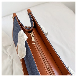 Detail of blue denim tote bag featuring brown leather trim and golden metal zipper closure