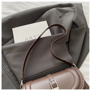 Brown leather saddle bag with silver buckle displayed on a gray blazer with books