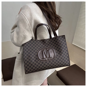 Detail of dark brown tote bag with M letter prints and shoulder strap in home setting