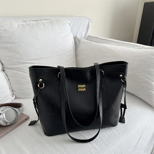 Minimalist black leather tote bag with gold logo and dual straps on a white sofa
