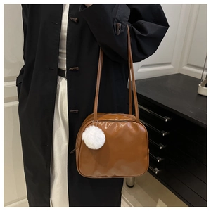 Brown faux leather shoulder bag with white pom-pom charm shown in a city style setting